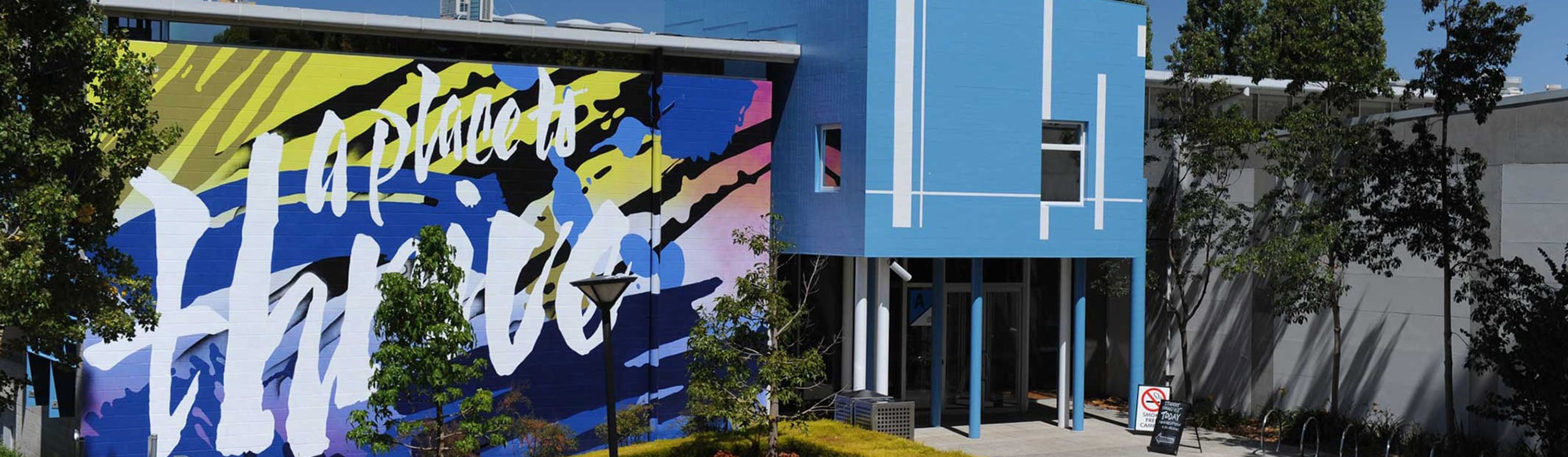 Facade of Greensborough campus. The entry is a blue building with a large mural that says "a place to thrive" on a blue, pink and yellow background